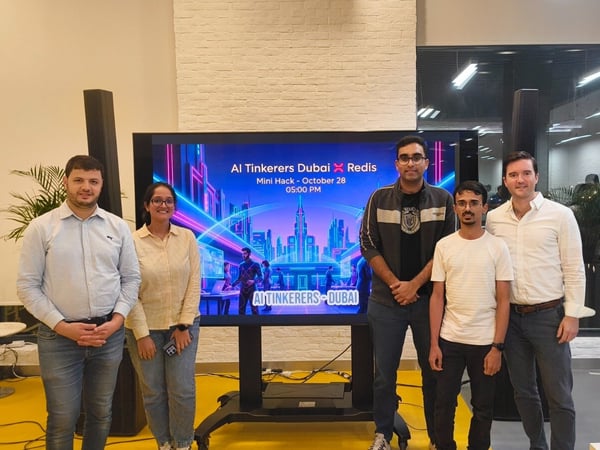 Five individuals, including four men and one woman, stand side-by-side posing for a group photo in an indoor setting, centered around a large digital screen advertising an 'AI Tinkers Dubai X Redis Mini Hack.'