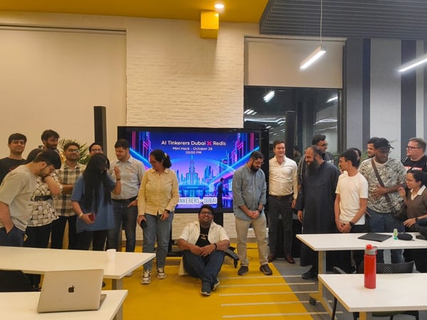 A group of approximately seventeen people are gathered indoors in a modern office or event space, posing for a photo in front of a large screen displaying event details for an AI Tinkerers Dubai X Redis Mini Hack.