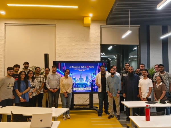 A group of approximately 18 people, likely attendees or organizers of a technical meetup, pose for a group photo indoors in front of a large monitor displaying the event title, "AI Tinkerers Dubai X Redis."