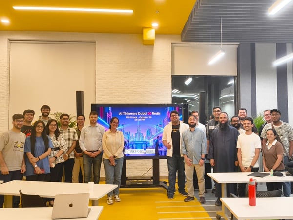 A group of approximately 16 individuals, mostly young adults, poses for a group photo inside a modern event space or office with bright yellow and white decor.