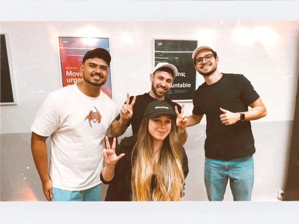 A group photo of four young adults, three men and one woman, posing indoors against a light-colored wall with posters, all smiling and making peace signs. The image has a vintage filter effect with a white border.