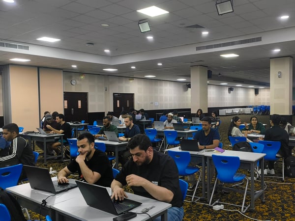A group of people, mostly young men, are seated at tables in a large, well-lit hall, working on their laptops.