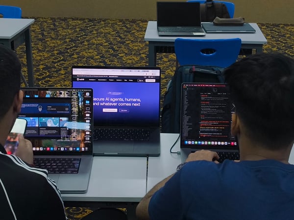 Two individuals are seated at a table, focused on their laptops in what appears to be a classroom or workspace setting.