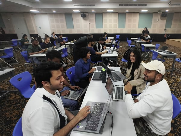 A group of people are seated at desks with laptops in a large hall, some working individually while others are engaged in conversation.