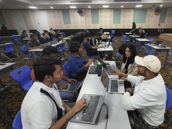 A group of young adults are gathered in a large hall, sitting at desks and working on laptops, likely during a workshop or educational event.