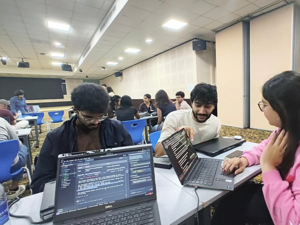 A group of young people are working collaboratively on laptops in a large, well-lit indoor hall, likely during a coding workshop or hackathon.