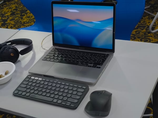 A workstation setup on a white table featuring an open MacBook Pro, an external Logitech keyboard, a mouse, and a pair of black headphones.