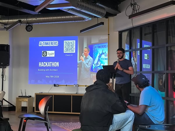 An indoor photograph showing a presenter speaking with a microphone next to a large projection screen displaying 'AI TINKERERS HACKATHON' and 'lyzr', with audience members seated in the foreground.