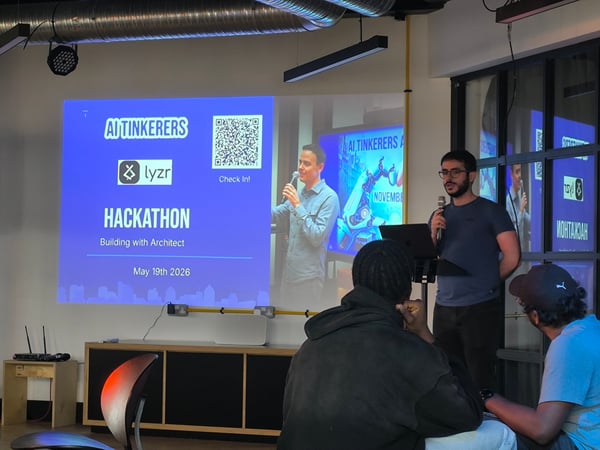 A man stands at the front of a room presenting at an AI Tinkerers Hackathon, with a large blue projection screen behind him displaying event details and a QR code.