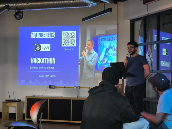 A presenter stands with a microphone next to a projector screen displaying information about an AI Tinkerers Hackathon, while audience members watch from the foreground.