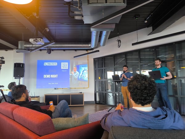 An indoor scene of an 'AI Tinkerers Demo Night' presentation, where two speakers stand near laptops facing an audience seated on couches in the foreground.
