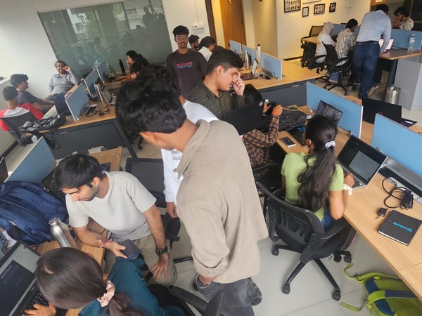 A wide-angle photograph of a busy indoor co-working space or office where several young adults are working on laptops at shared desks with blue partitions.