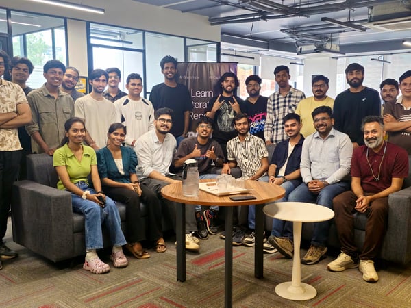 A large group of young adults, mostly men, posing for a group photo in a modern office setting with sofas and a banner in the background.