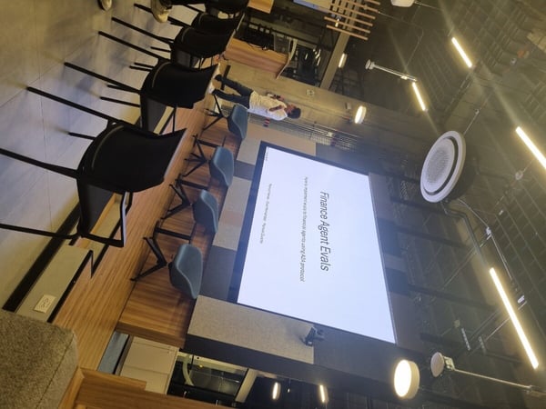A photograph of a presentation titled 'Finance Agent Evals' displayed on a large screen in a modern office or event space with rows of empty chairs.