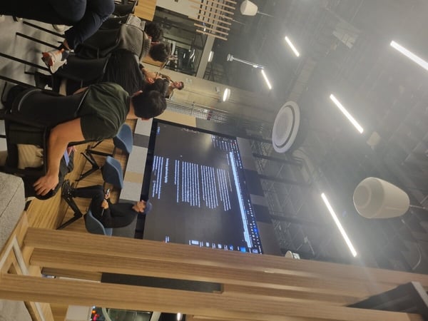 A group of people are seated in a modern, industrial-style indoor space, watching a presentation displayed on a large screen.