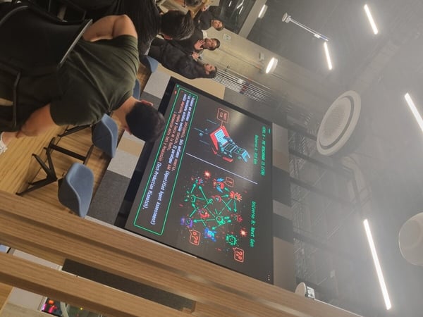 A high-angle, slightly tilted photograph of a group of people attending a presentation in a modern indoor setting, featuring a large screen with Spanish text about AI benchmarks.