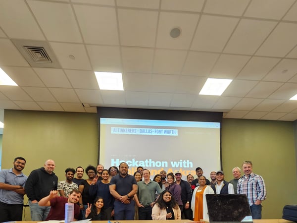 A group of people posing for a group photo in a conference room in front of a projector screen announcing an AI Tinkerer's Hackathon.