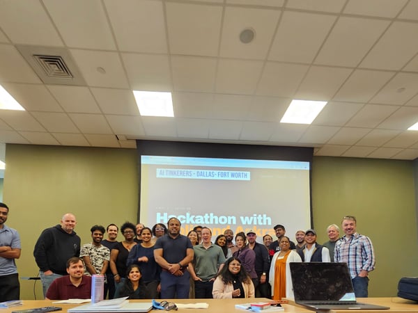 A group of people posing for a commemorative photo in front of a projection screen during an AI Tinkerers hackathon event.