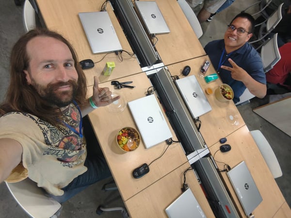 A top-down selfie of two men sitting at a long wooden desk filled with HP laptops, enjoying a meal in a workspace environment.