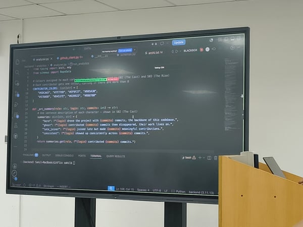A photograph of a large display screen in a room showing Python code in a text editor, with a person standing next to a wooden podium on the right.