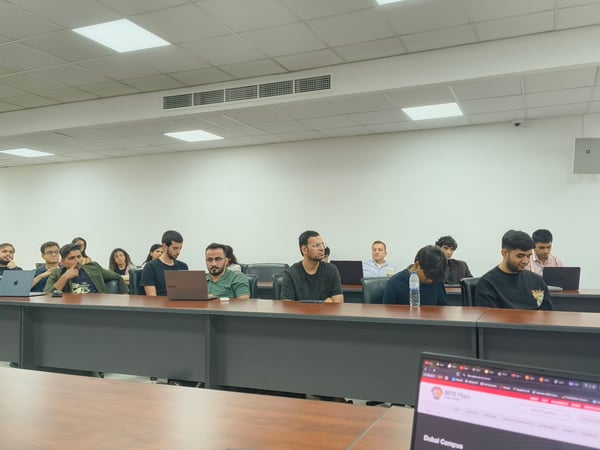 An indoor photograph showing an audience of students or professionals seated at long rows of desks in a classroom or lecture hall, with a laptop in the foreground displaying a webpage.