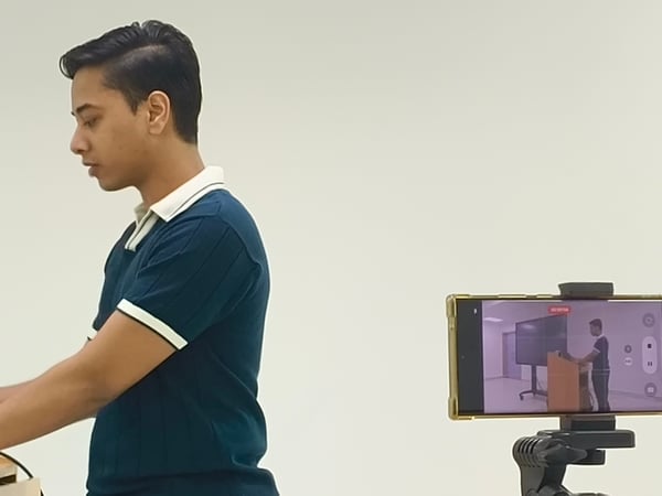 A young man in a dark blue polo shirt stands at a podium in profile, while a smartphone mounted on a tripod in the foreground records his presentation.