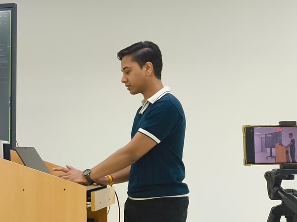 A man stands at a wooden podium using a laptop while being recorded by a smartphone mounted on a tripod.
