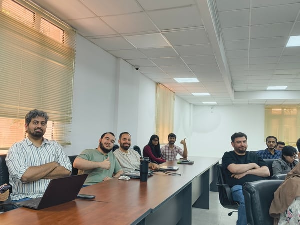 A group of colleagues or students are sitting around a long wooden conference table in a bright office or classroom setting, with several of them looking towards the camera.