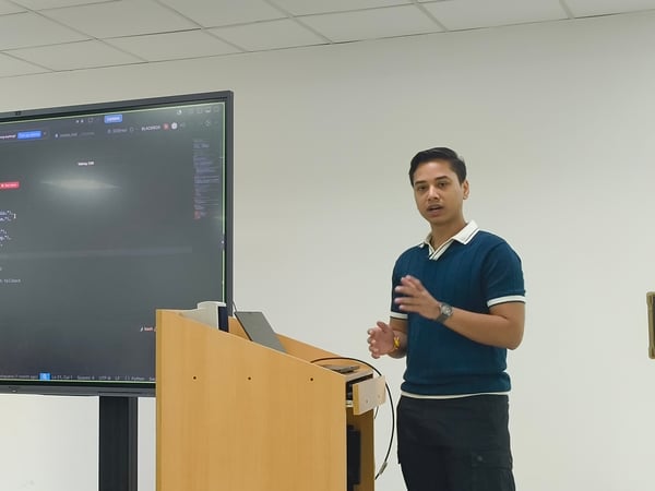 A young man stands next to a large digital display screen showing Python code, delivering a presentation or demonstration in an indoor setting.
