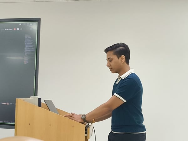 A young man stands at a wooden podium, typing on a laptop while presenting. Behind him, a large digital screen displays code and text.