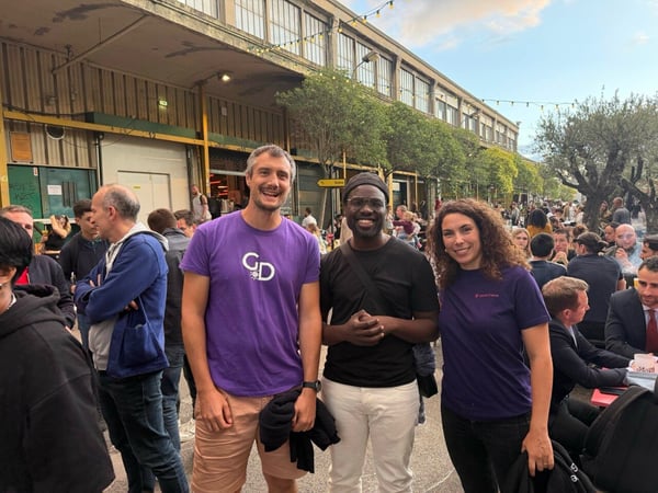 A photo of three people—two men and one woman—smiling at the camera during a crowded outdoor social event or networking gathering, set against a backdrop of an industrial-style building and trees with string lights.