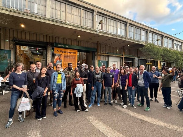 A large group of approximately thirty people poses for a casual group photo outdoors in front of a long, industrial-style building facade featuring an orange sign for "Food-express."