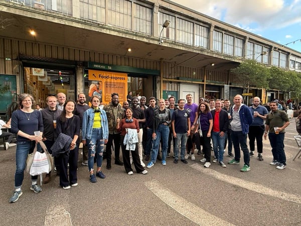 A large group of approximately 28 people, likely colleagues or friends, poses for a group photo outdoors in front of a long, industrial-style building with large windows and a covered market area.