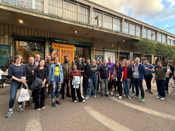 A large group of approximately 27 people, dressed casually, stand together outdoors for a group photo in front of a long, industrial-style building facade that features a "Food-express" entrance.