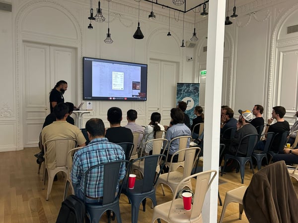 A photograph capturing an indoor presentation where a man addresses an audience seated in rows of metal chairs, with a large screen displaying technical content behind him. The event is taking place in an ornate room with high white walls and decorative molding.