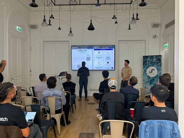 A photograph capturing an indoor event where a group of people is attending a presentation in an ornate white room with high ceilings. Two speakers stand near a large screen displaying the presentation content, while the audience is seated facing them.