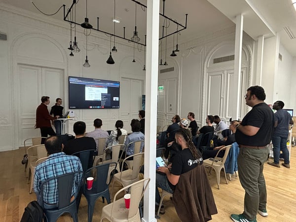 A photograph capturing a technical presentation or meetup taking place indoors in a large, ornate room with high ceilings and industrial lighting fixtures. Two presenters stand near a large screen displaying code or a dashboard, addressing an audience seated in rows of chairs.