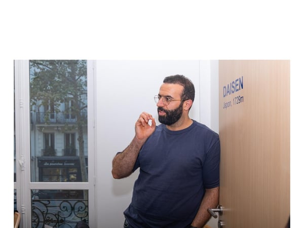 A candid indoor photograph of a bearded man wearing glasses and a dark blue t-shirt, standing next to a light wooden door and a large window overlooking a city street scene.