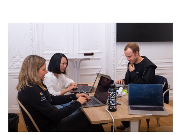 A candid indoor photograph shows three people (two women and one man) sitting at a table, working intently on their open laptops in a bright room with ornate white wall molding.