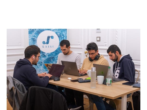 A candid photograph shows four men sitting closely around a wooden table in an indoor setting, working on their laptops in what appears to be a focused work or coding session. A blue banner featuring a logo and the word 'RAISE' is displayed on the ornate white wall behind them.