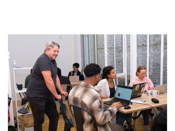A group of young adults works on laptops at a table indoors while a standing man leans over a chair and observes them with a smile, likely in a collaborative office or workshop setting.