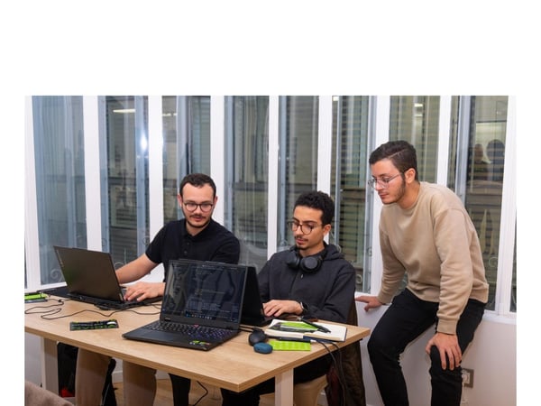 Three young men are collaborating on code displayed on laptops at a wooden desk in a bright indoor setting, likely an office or co-working space. Two are seated and one is standing, all focusing on the screens.