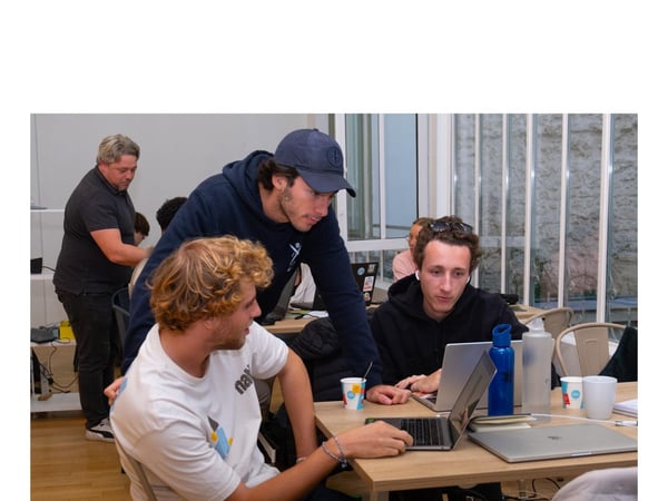 A candid photograph showing a group of young adults gathered around a table indoors, focusing on open laptops during a collaborative work session or meeting.