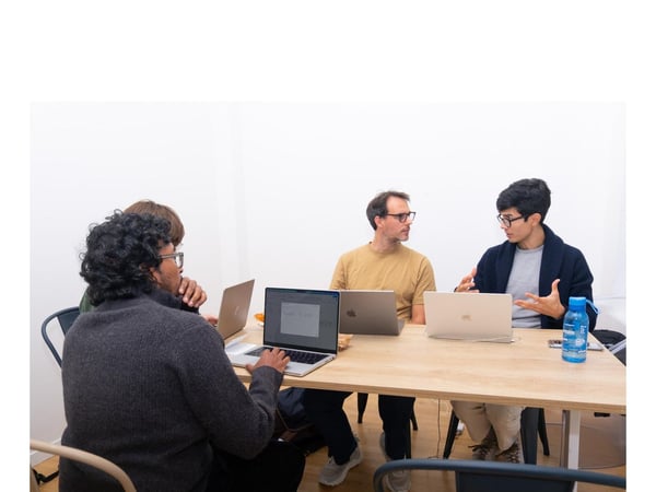 Four individuals are seated around a light wooden table in a bright, minimalist room, engaged in a collaborative discussion while using open laptops.