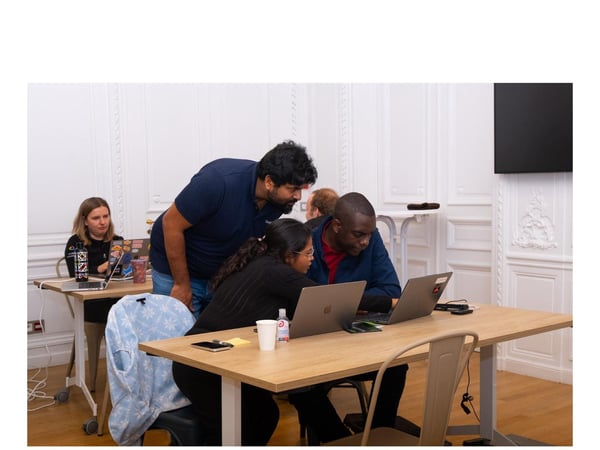 A candid photograph captures three people collaborating closely, looking at content on two laptops on a wooden table in a brightly lit, ornate meeting room.