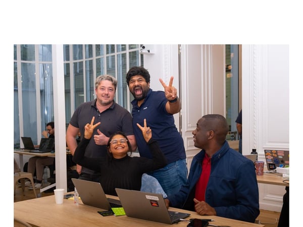 A candid photograph captures a diverse group of four people posing happily around a wooden table with laptops, with three of them making the peace sign, in an indoor office or event setting.