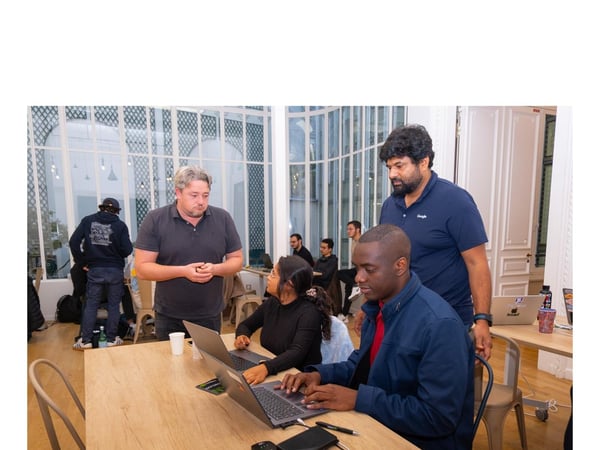 A candid photograph capturing a group of people collaborating around laptops at a large wooden table in a brightly lit indoor space with large, arched glass windows in the background.