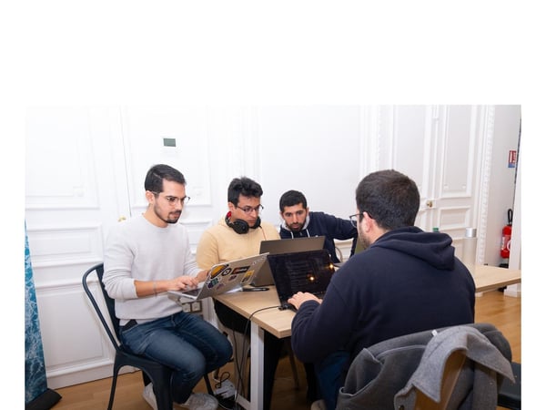 Four men are seated around a light wooden table in a brightly lit office setting, focused on working on their individual laptops. They appear to be engaged in a collaborative coding or work session.