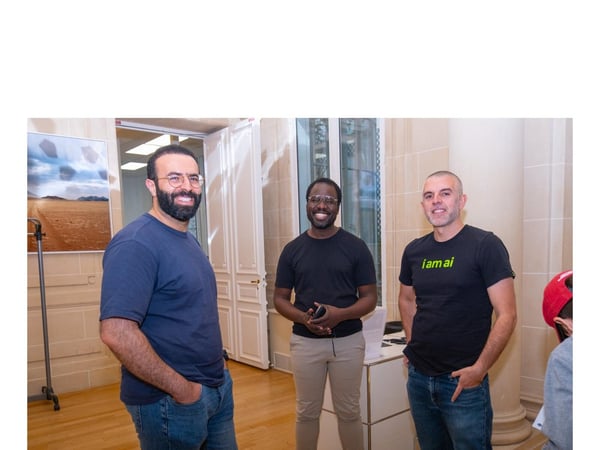A photograph captures three men standing and smiling in a well-lit, formal indoor setting, possibly at an event, with one of the men wearing a black t-shirt that reads "i am ai."
