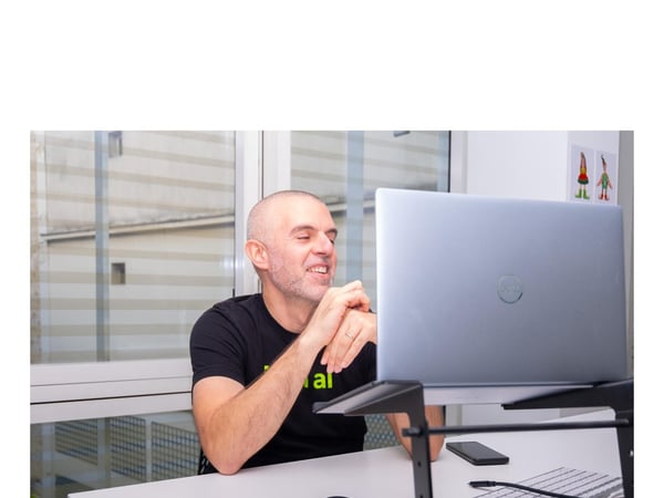 A man with a shaved head is seated at a white desk indoors, smiling and looking intently at a silver laptop screen placed on a black adjustable stand, suggesting he is participating in a video conference or remote work session.